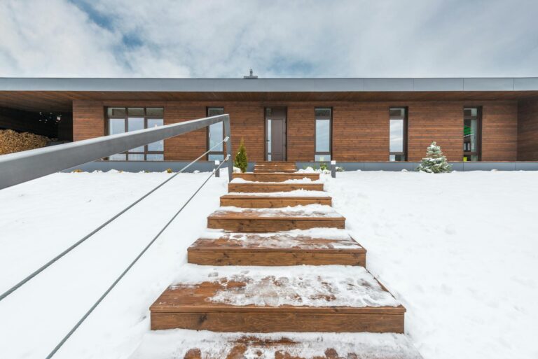 A contemporary wooden house with snow-covered steps leading to the entrance, captured in a winter setting.